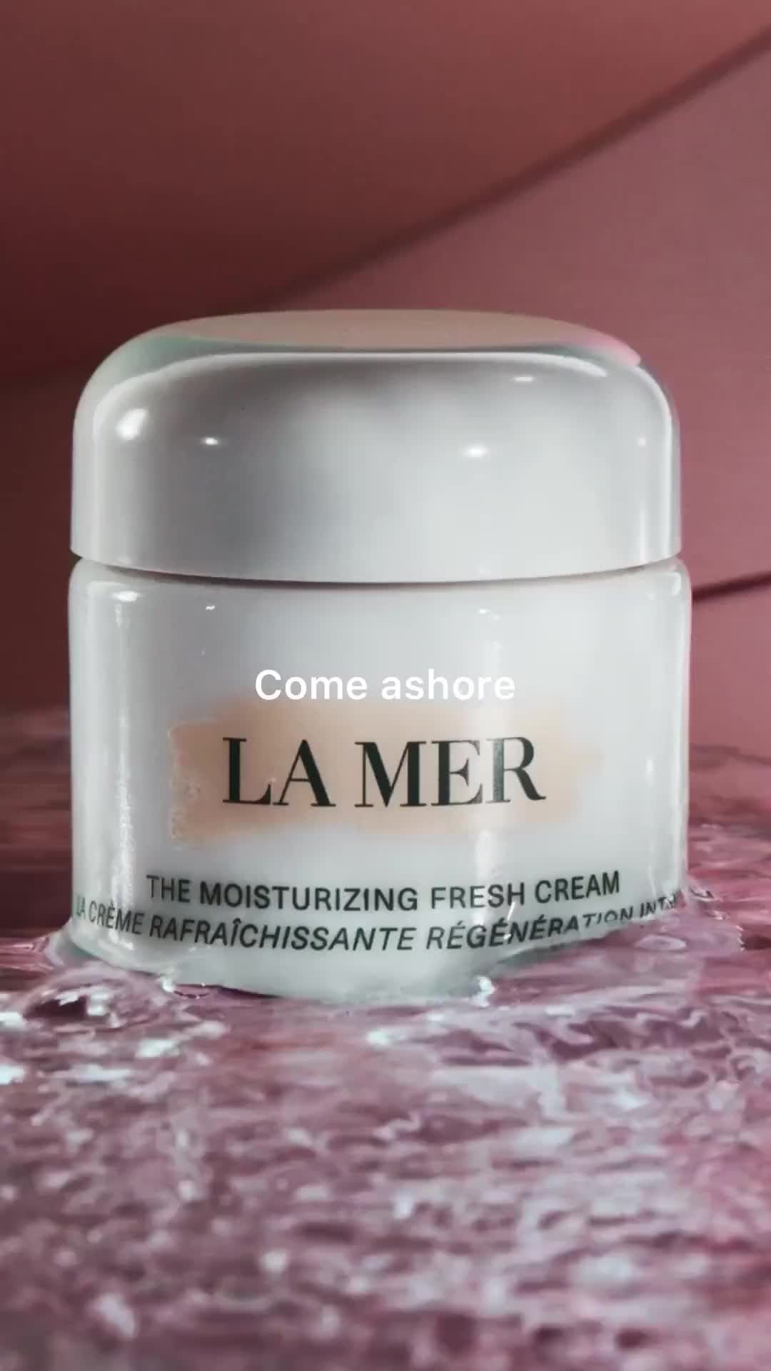 Close-up of a white La Mer moisturizing cream jar on a reflective pink surface. Product shot with a pink background.