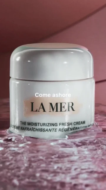 Close-up of a white La Mer moisturizing cream jar on a reflective pink surface. Product shot with a pink background.