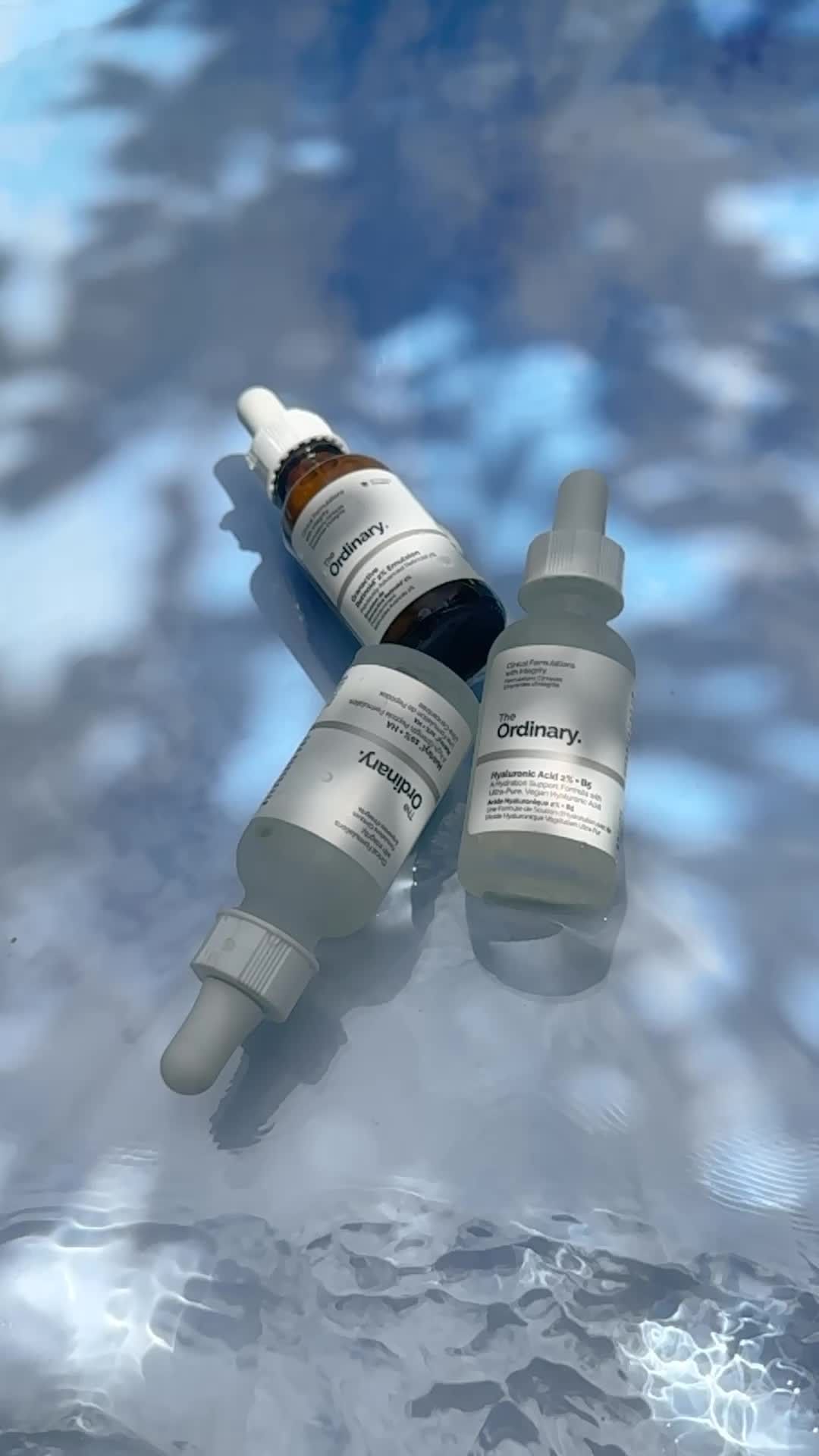 Three The Ordinary skincare bottles are arranged on a rippled water surface with a blue and white sky reflection for a beauty product shot.