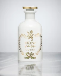 An opaque white ceramic perfume bottle with gold and green illustrations resting on a reflective surface