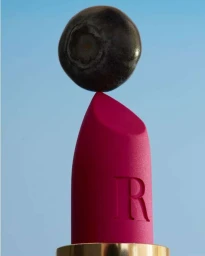 A single dark blueberry balanced precariously on the sharp point of a matte magenta lipstick against a clear blue background.