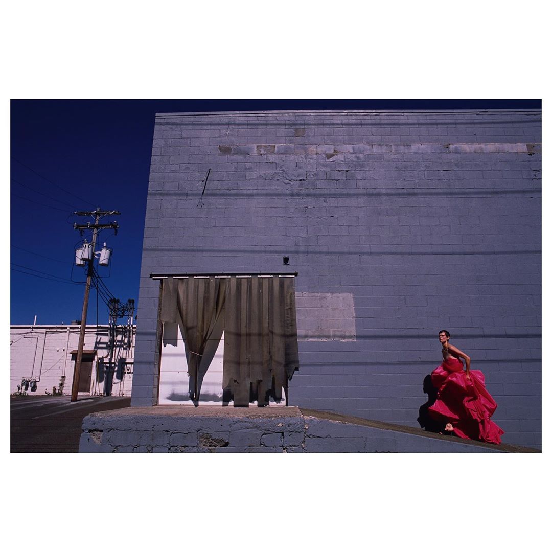 Woman in red dress stands on loading dock ramp against a grey brick building under a deep blue sky in an outdoor fashion shot.