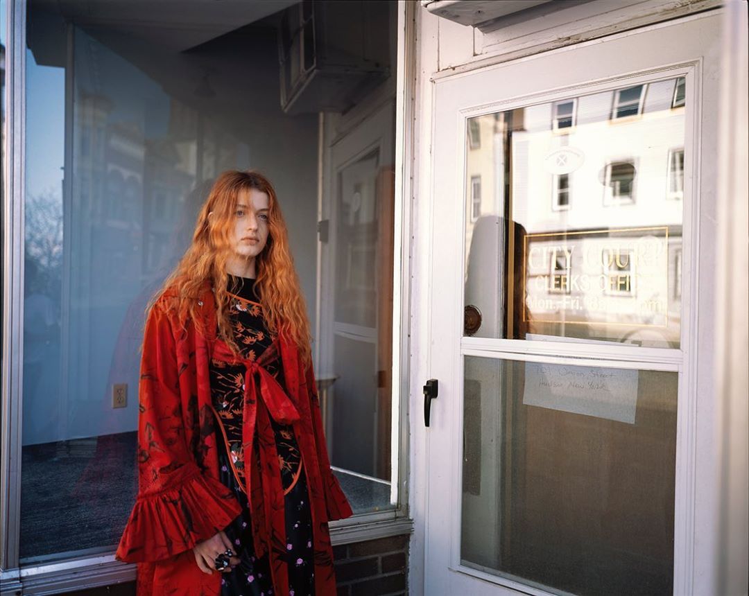 Woman with red hair in front of a door wearing a red floral robe over a black dress, standing outside on a sunny day.