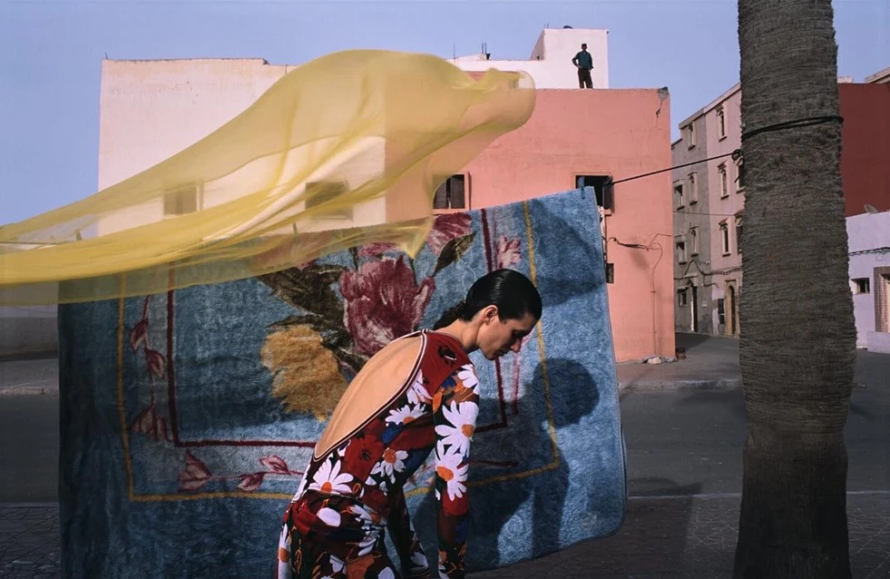 Fashion shot of a woman in floral dress, standing in a city street near a palm tree, carpet, and flowing yellow textile in daytime.