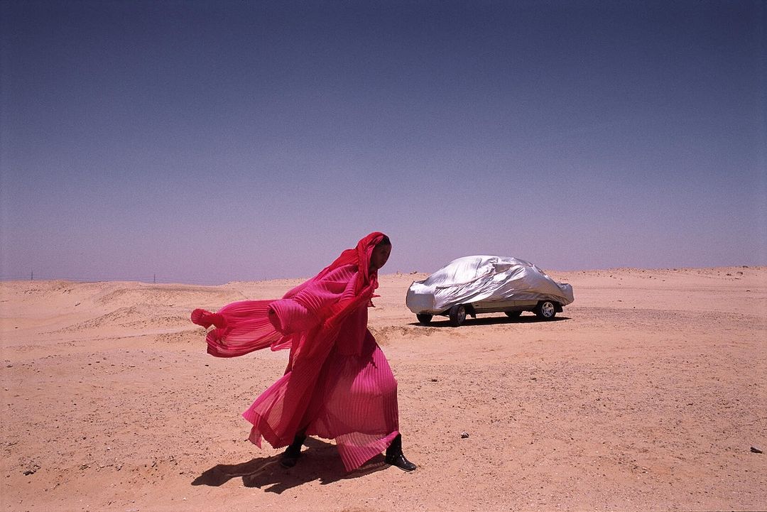 Woman in red dress walks in desert near car under silver cover on sunny day with clear blue sky. Fashion editorial shot.