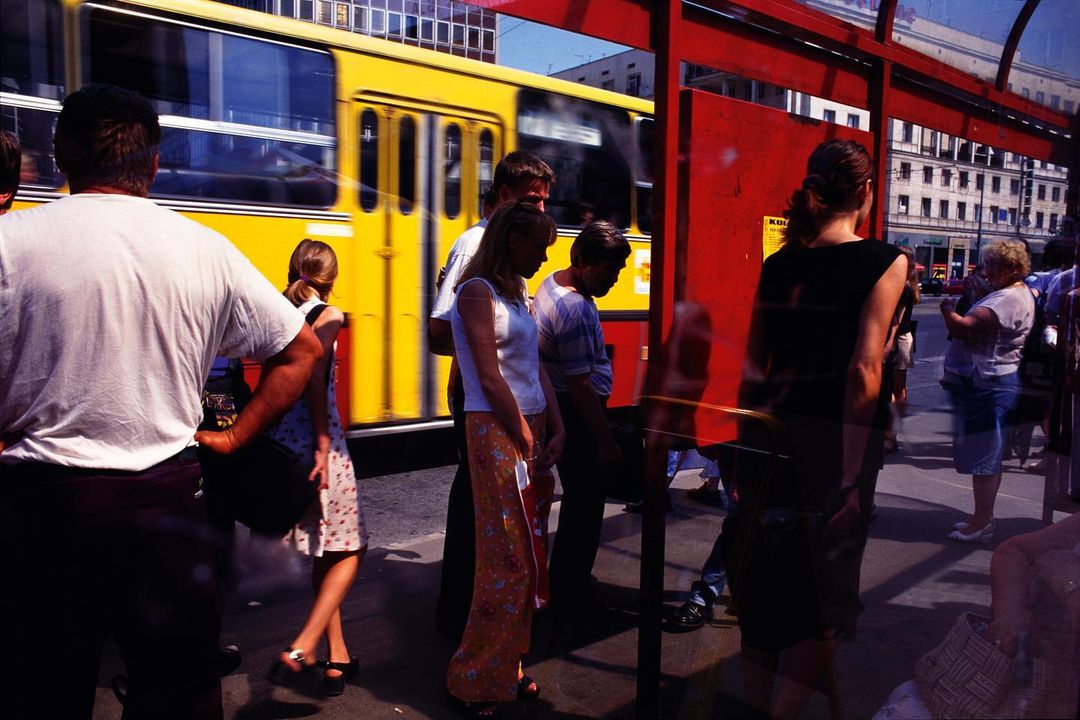 People wait at a bus stop as a yellow bus drives by in an urban environment on a sunny day, with buildings visible in the background.