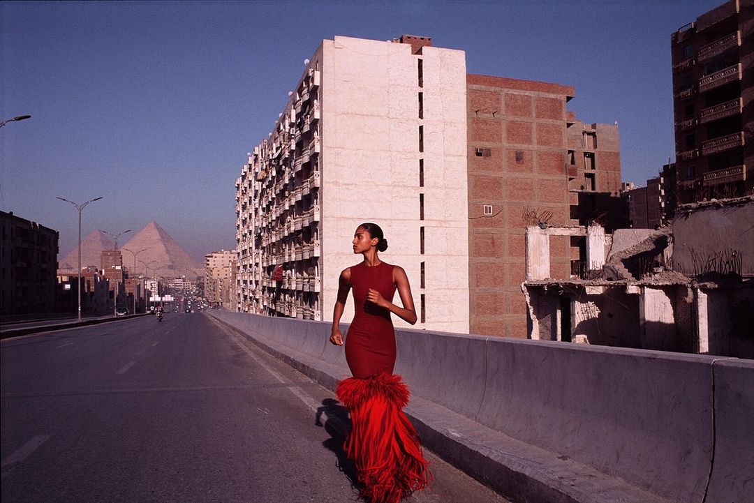 Woman in red dress runs on an elevated road with buildings and pyramids in the background, creating a striking fashion editorial image.