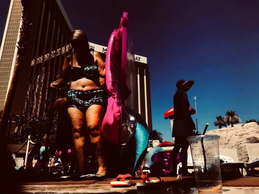 Woman in swimwear stands near a pool with a pink flamingo inflatable. Another woman is walking in the background near resort architecture and palm trees.