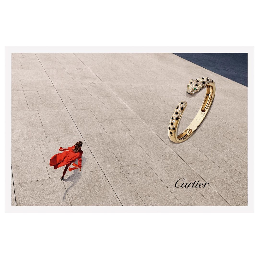 High-angle view of a Cartier gold bangle bracelet with jaguar heads, contrasted by a distant woman in a red coat on a concrete plaza.