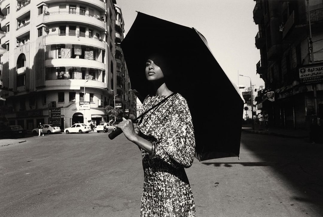 Woman in patterned dress holding a large black umbrella on a city street by Vincent Van De Wijngaard