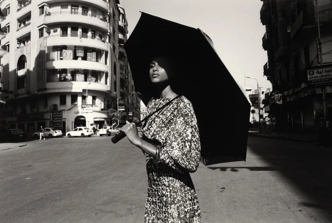 Woman in patterned dress holding a large black umbrella on a city street by Vincent Van De Wijngaard
