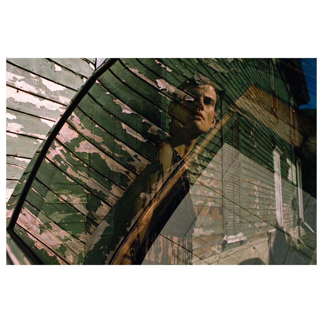 An adult male stands near a weathered green wooden building, creating a layered, textured visual with a unique perspective and aesthetic.