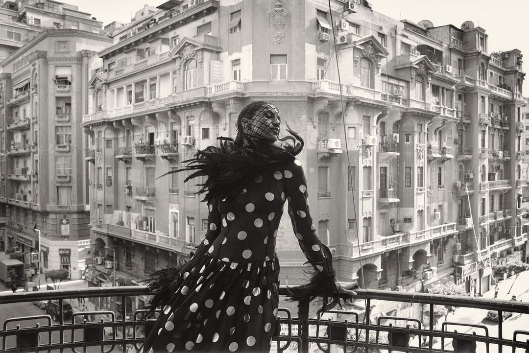 Monochrome fashion portrait of an adult woman in a polka-dot dress and feathered accents posing on a balcony overlooking a city street.