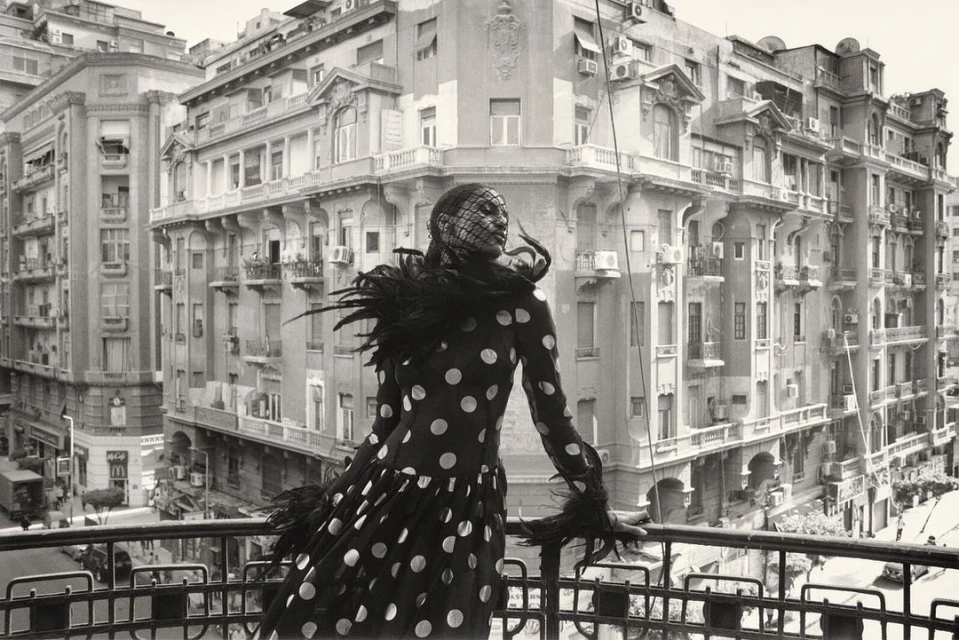 Monochrome fashion portrait of an adult woman in a polka-dot dress and feathered accents posing on a balcony overlooking a city street.