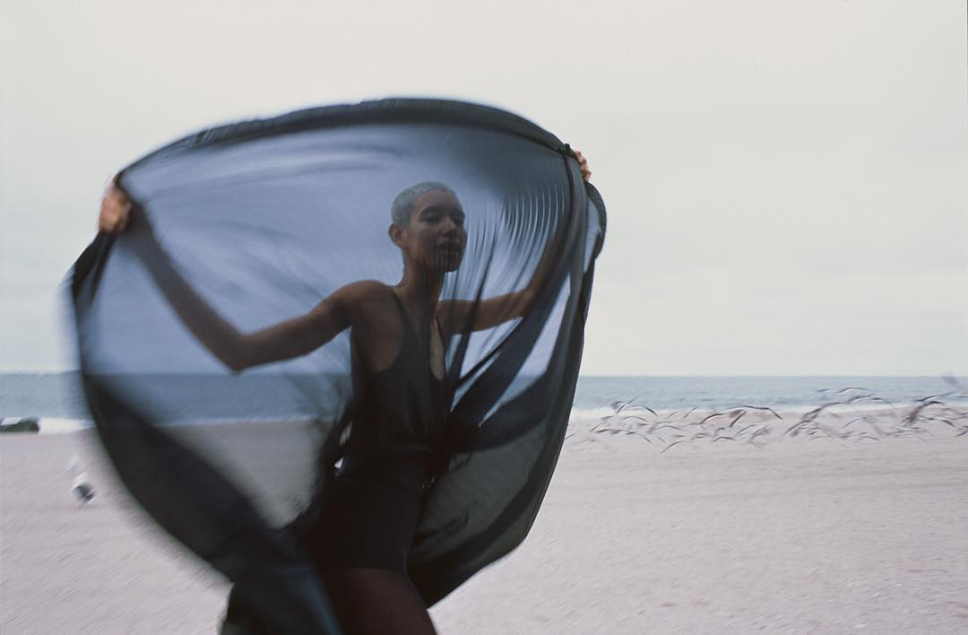 Woman in swimsuit and flowing dark fabric poses on a sandy beach with ocean and birds in background on an overcast day.