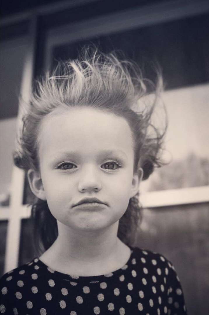 Black and white portrait of a young girl with windblown hair wearing a polka dot shirt, with a window frame visible in the background.