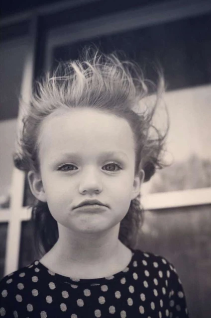 Black and white portrait of a young girl with windblown hair wearing a polka dot shirt, with a window frame visible in the background.