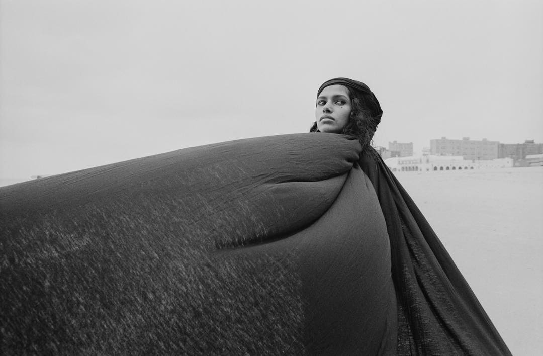 Monochrome portrait of a woman with dark hair and a headscarf, enveloped in a flowing robe with a building in the background.