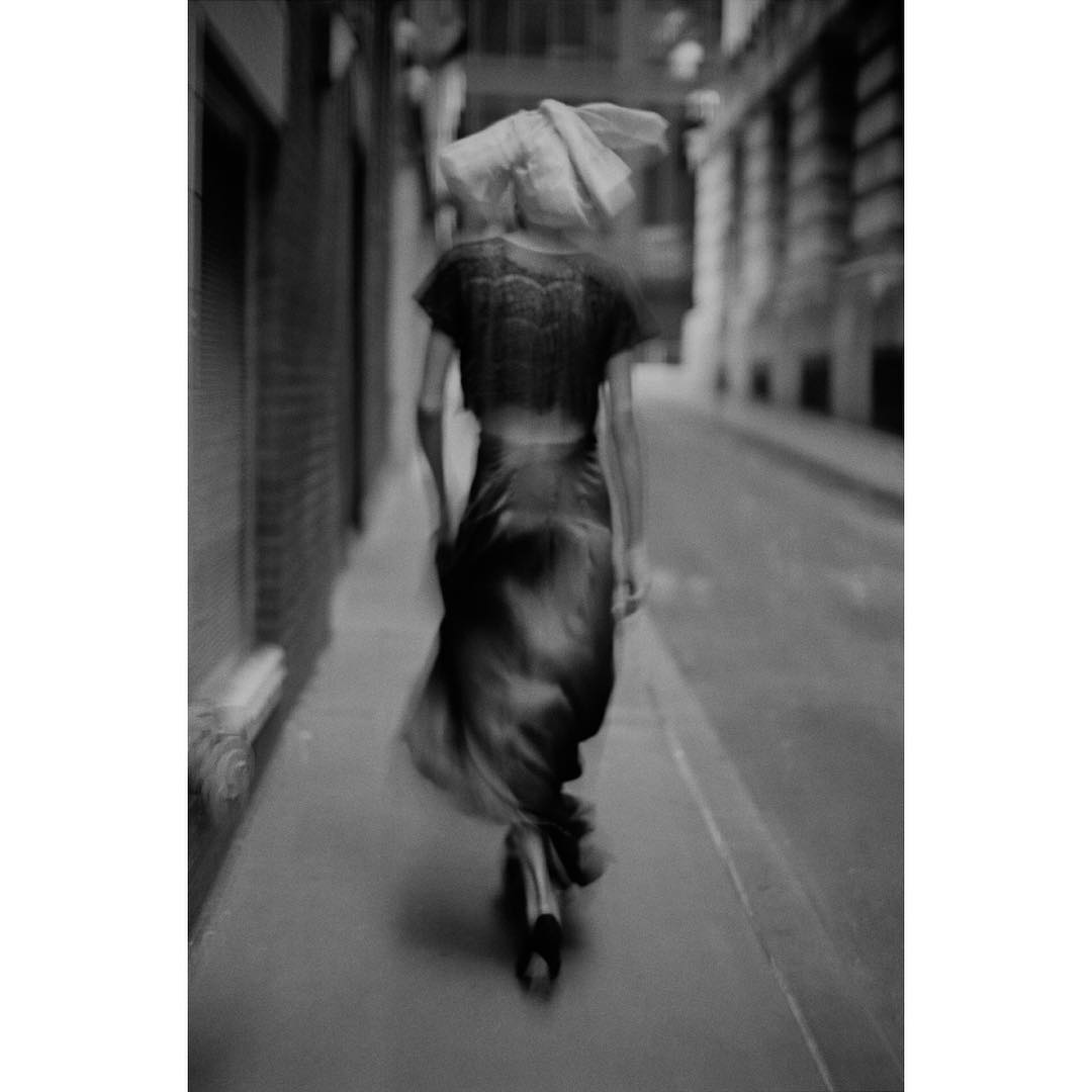 Monochrome motion blur of a woman walking down a city street, wearing a dress and hat, with blurred motion suggesting movement.