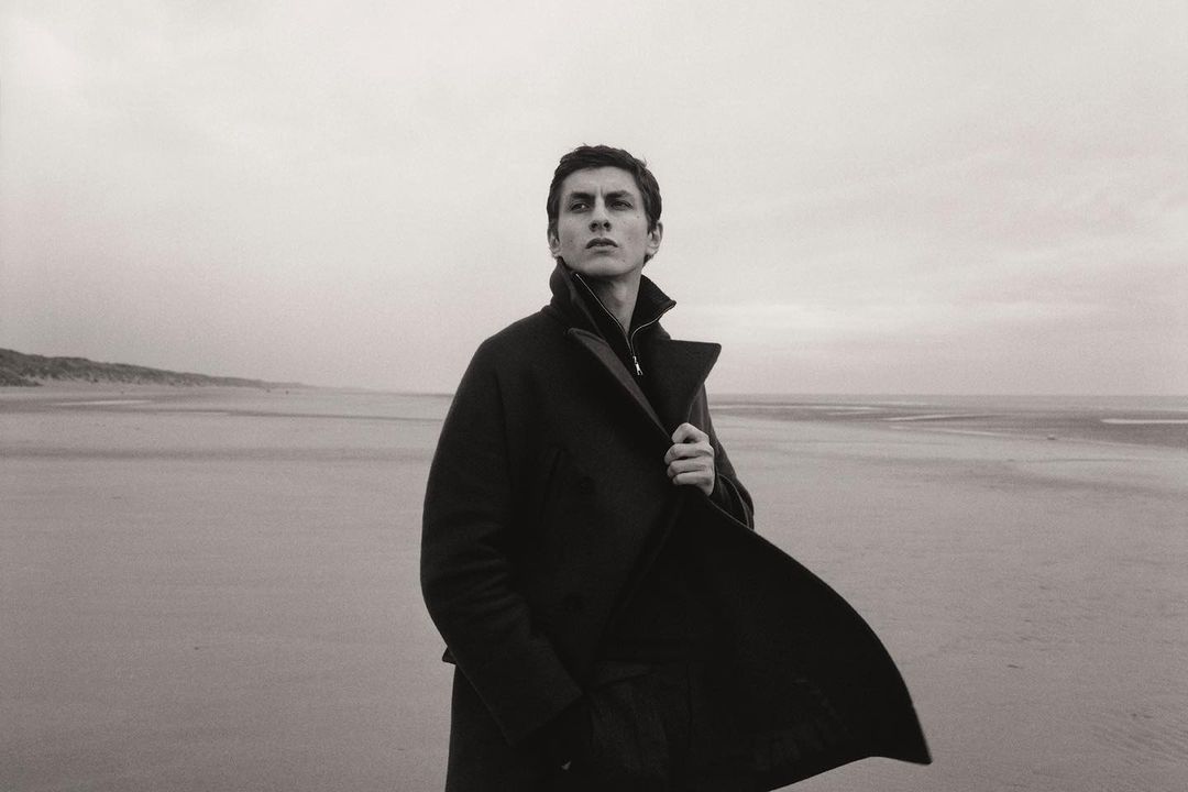 A man in a dark coat stands on a beach with a cloudy sky. He looks off into the distance, capturing a contemplative mood in monochrome.