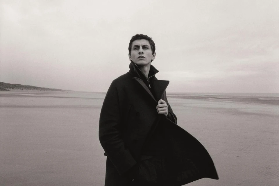 A man in a dark coat stands on a beach with a cloudy sky. He looks off into the distance, capturing a contemplative mood in monochrome.