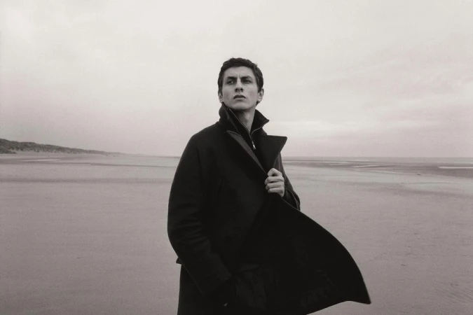 A man in a dark coat stands on a beach with a cloudy sky. He looks off into the distance, capturing a contemplative mood in monochrome.