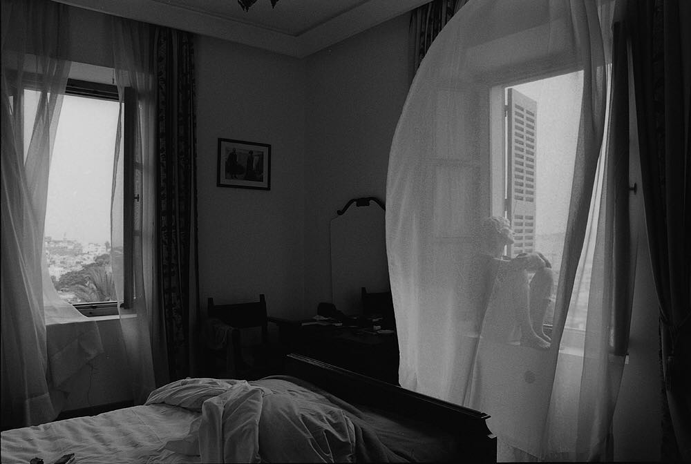 Black and white indoor scene of a child sitting in a window with sheer curtains, a bed, chair, and cityscape visible through the window.
