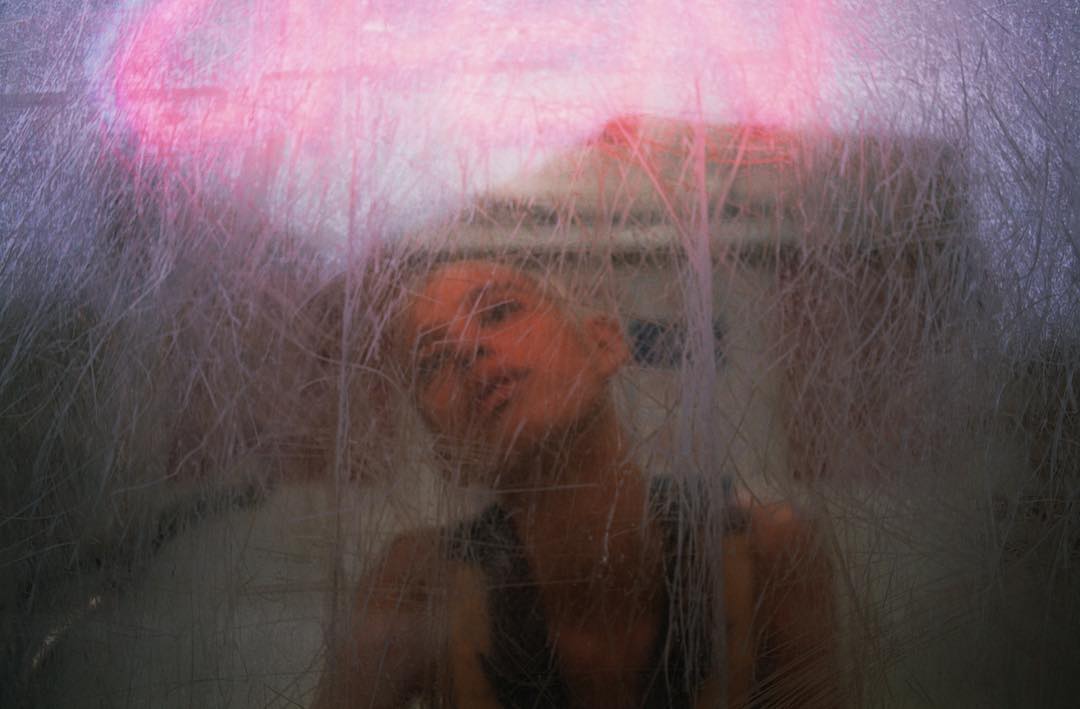 Abstract portrait of a person partially obscured by scratched glass or a textured surface with pink and white light in the background.