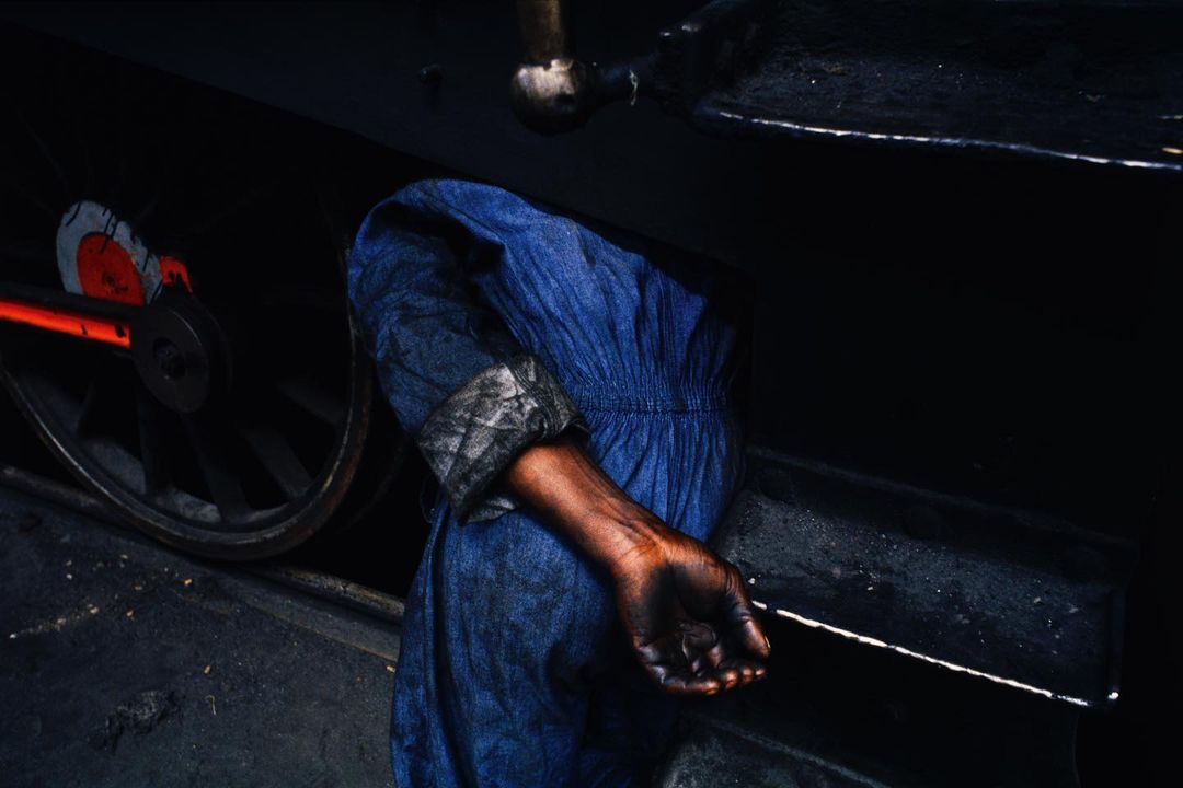 Close-up of a person in blue work clothes repairing a dark-colored train wheel; only the arm, hand, and sleeve are visible in the frame.