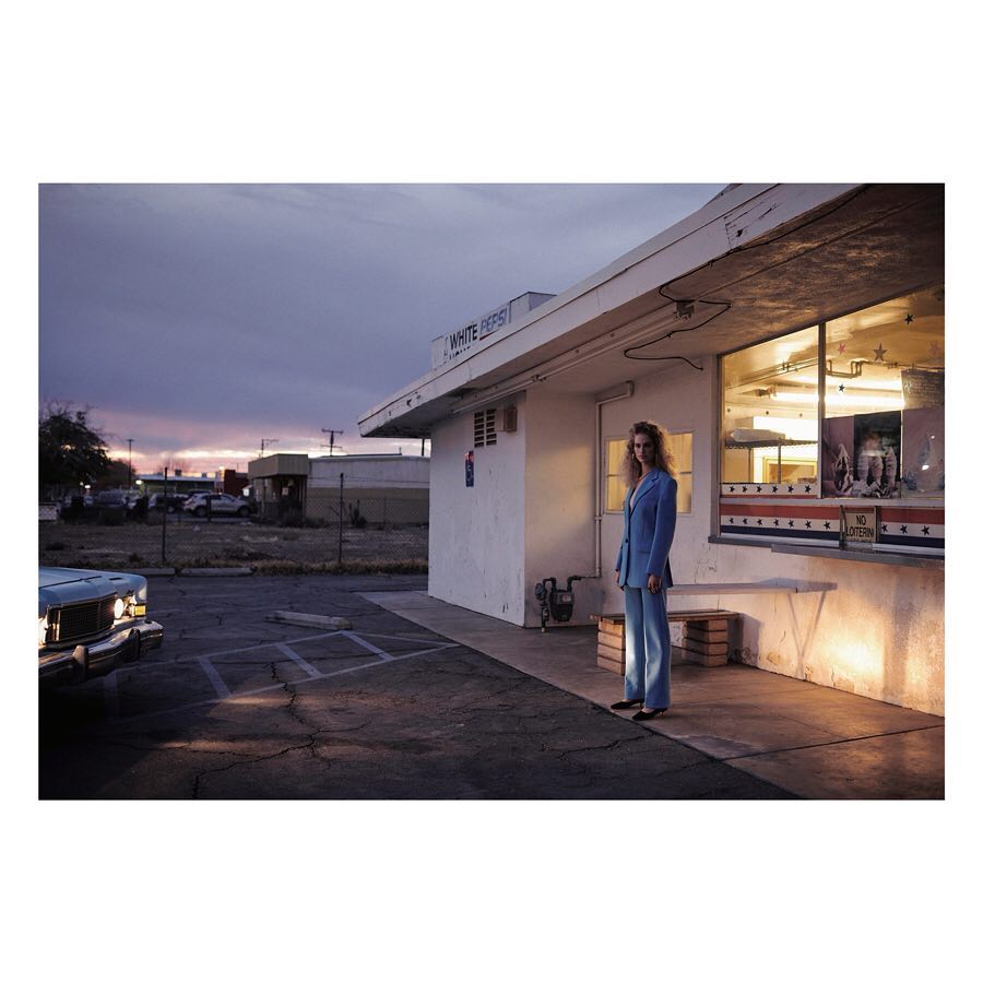 Woman in blue suit stands outside vintage diner at dusk. Classic car parked nearby, creating a retro, cinematic atmosphere.