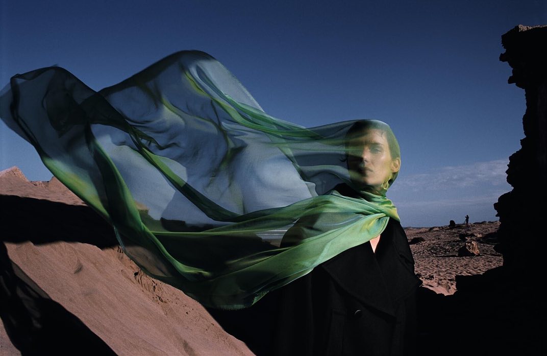 Model in a black coat with a flowing green sheer scarf whipping in the wind against a desert landscape