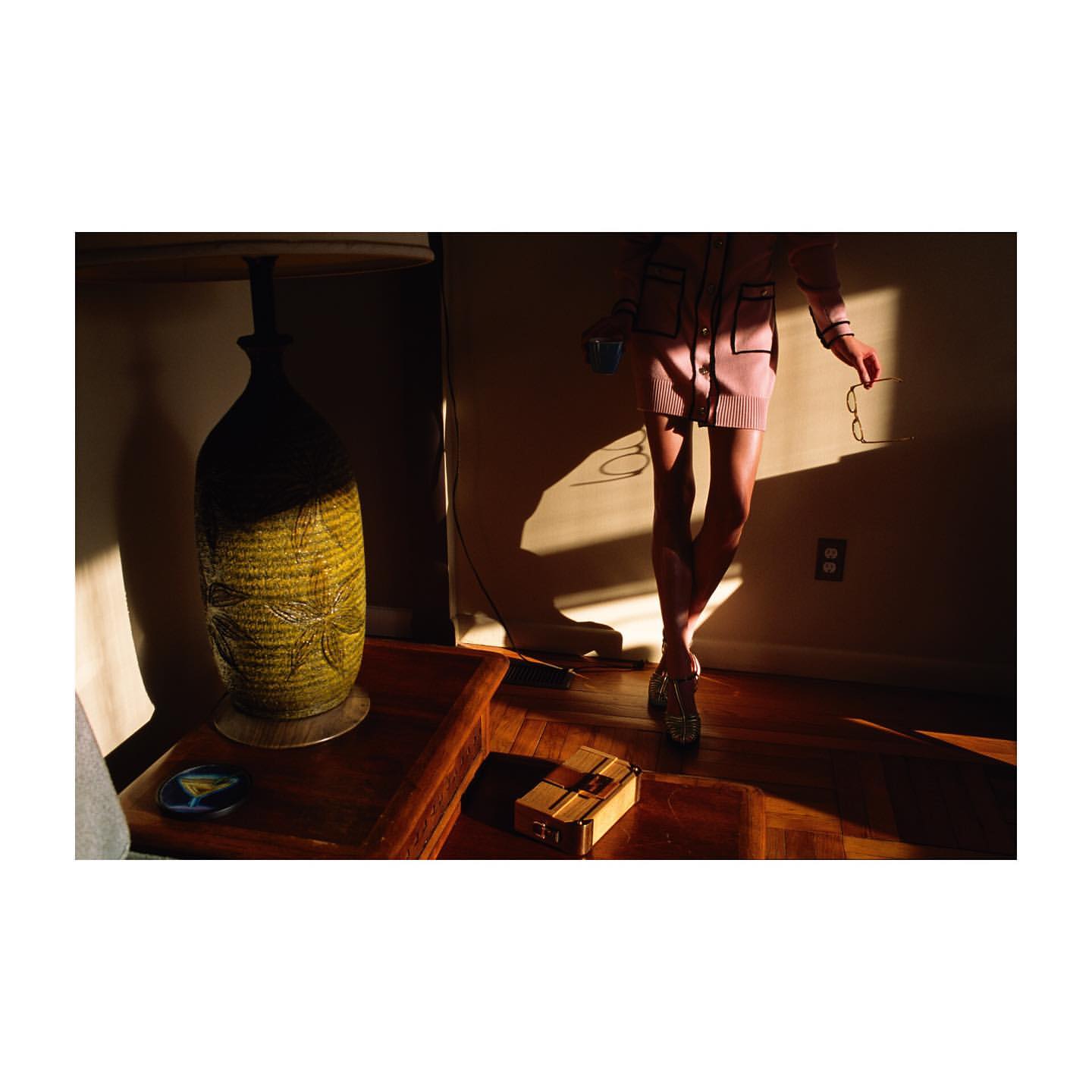 Woman in pink suit, indoors. Decorative lamp casts light on wood floor, briefcase, and wall. Shadows add contrast and depth.
