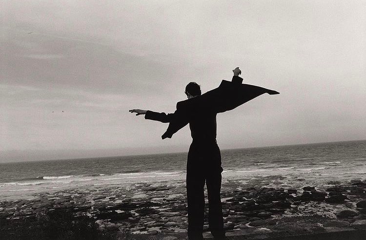 Person in a black suit stands on a rocky shoreline, facing the ocean, holding their jacket out to the sides, in black and white.