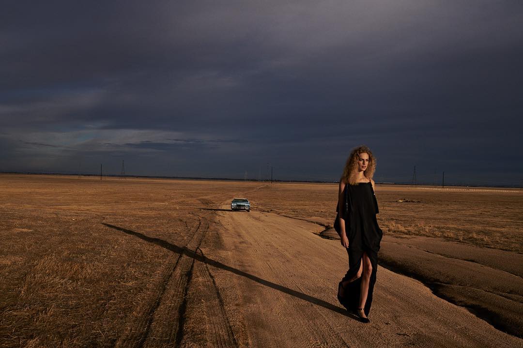 A woman in a black dress walks along a dirt road with a classic car in the distance against a dramatic sky in an outdoor setting.
