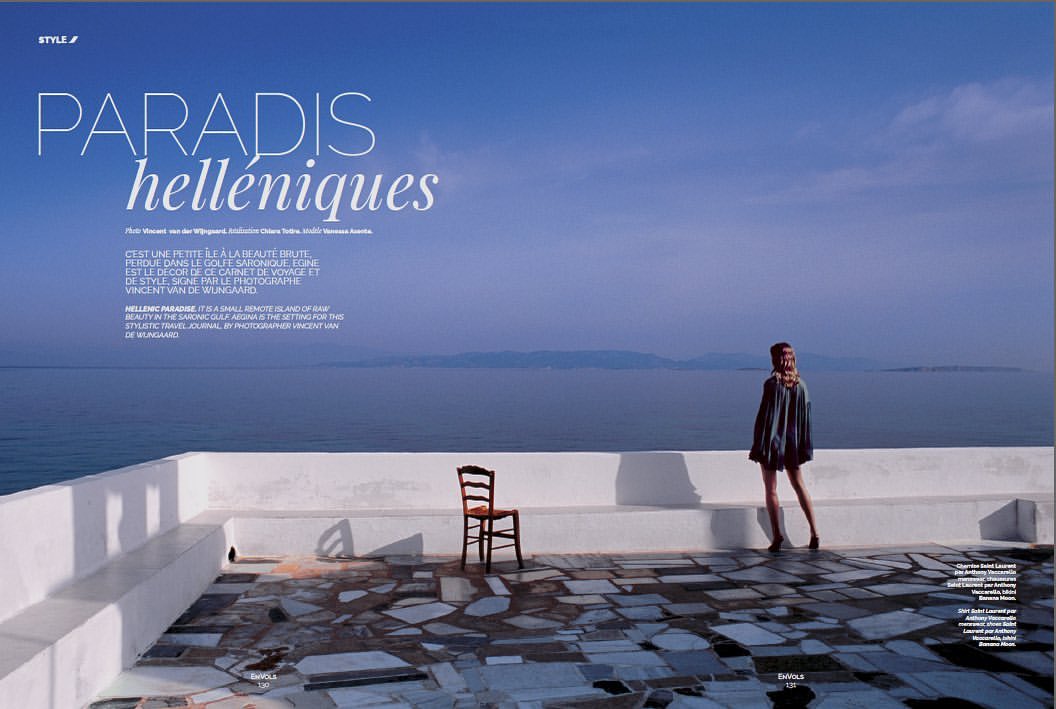 Woman poses on rooftop terrace with ocean view, wearing black tunic and hat; chair stands nearby against the white balcony wall.
