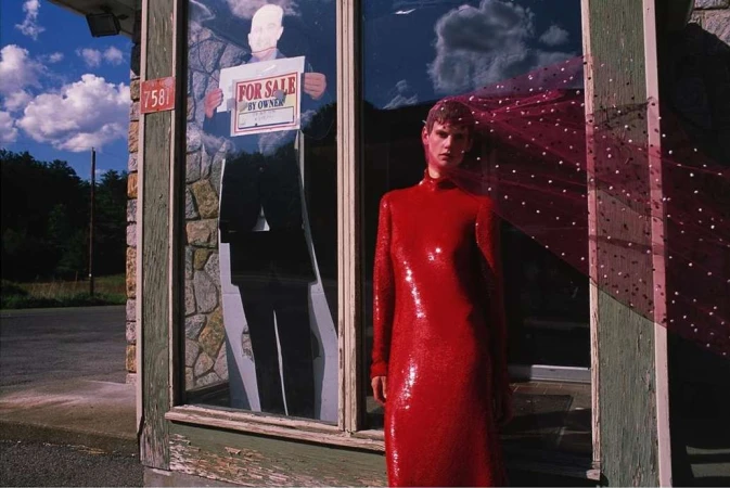 Adult female in sequined red gown posing outside abandoned stone building with a 'for sale by owner' sign visible in the window.