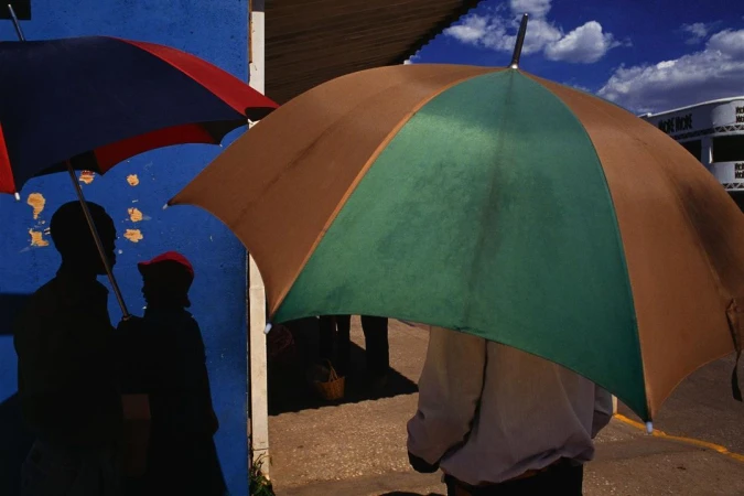 Silhouetted figures holding a blue and red umbrella alongside a person holding a large brown and green umbrella outdoors