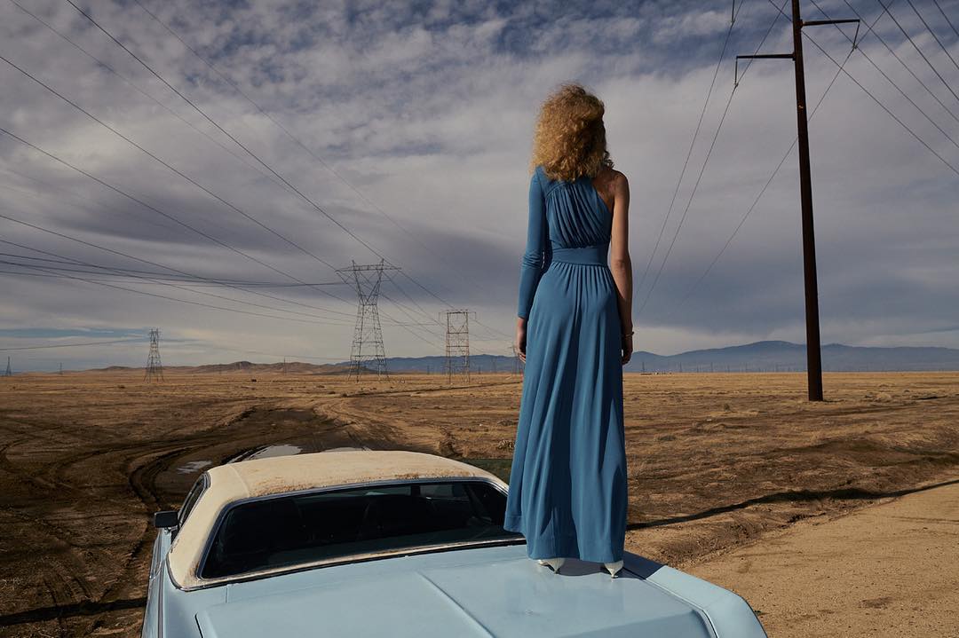 Woman in a blue gown standing on the trunk of a light blue car in a desert landscape under a cloudy sky with telephone poles.