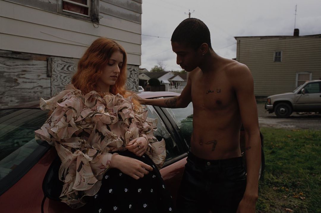 A red-haired woman in a floral blouse leans against a car, while a shirtless man with tattoos stands beside her in an outdoor scene.