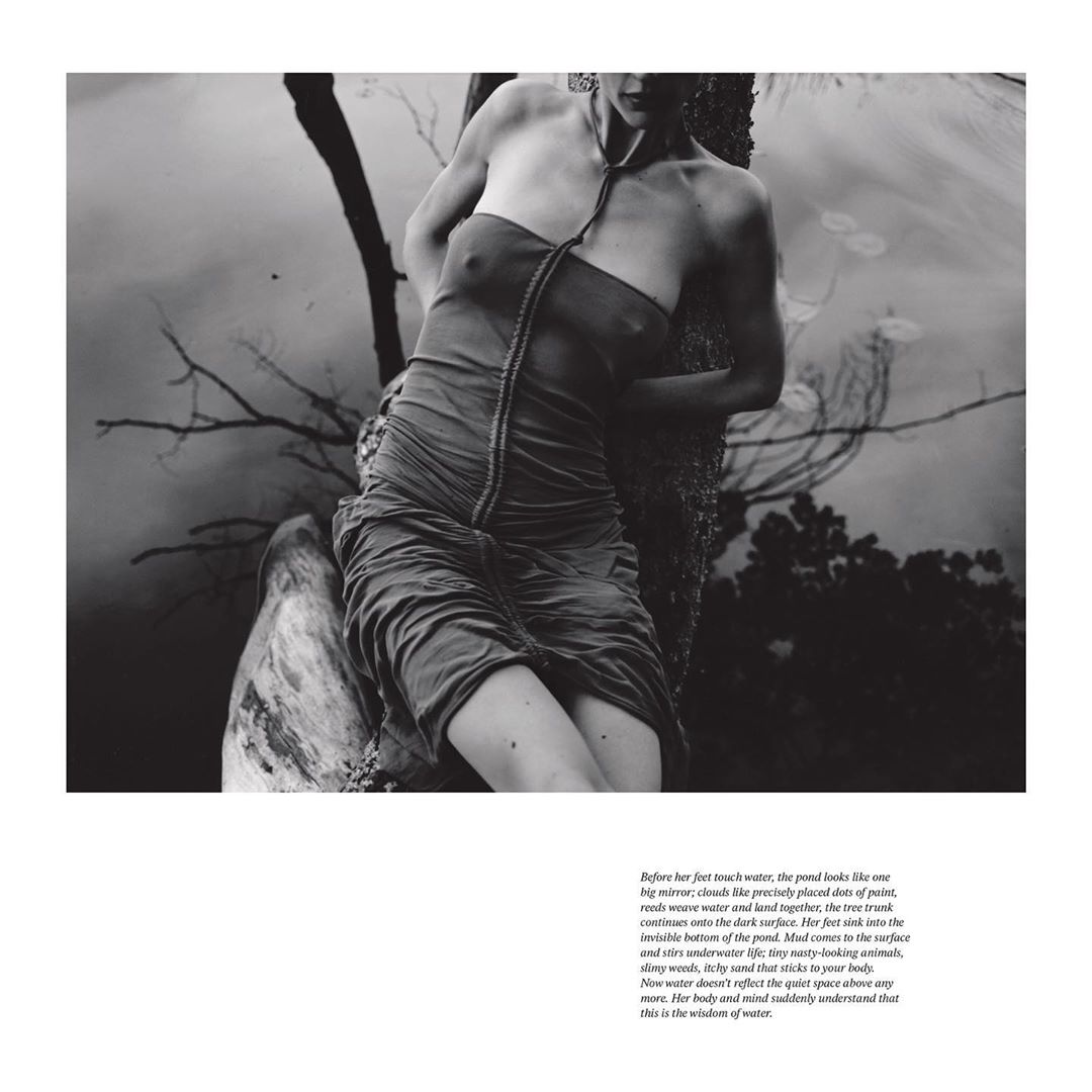 Monochrome portrait of a woman in a draped dress, partially submerged in water, leaning against a tree near a pond, in a fine art style.