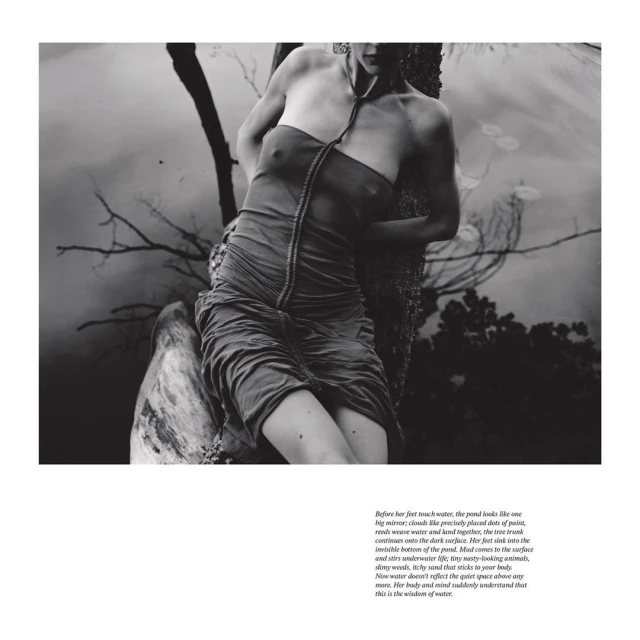 Monochrome portrait of a woman in a draped dress, partially submerged in water, leaning against a tree near a pond, in a fine art style.