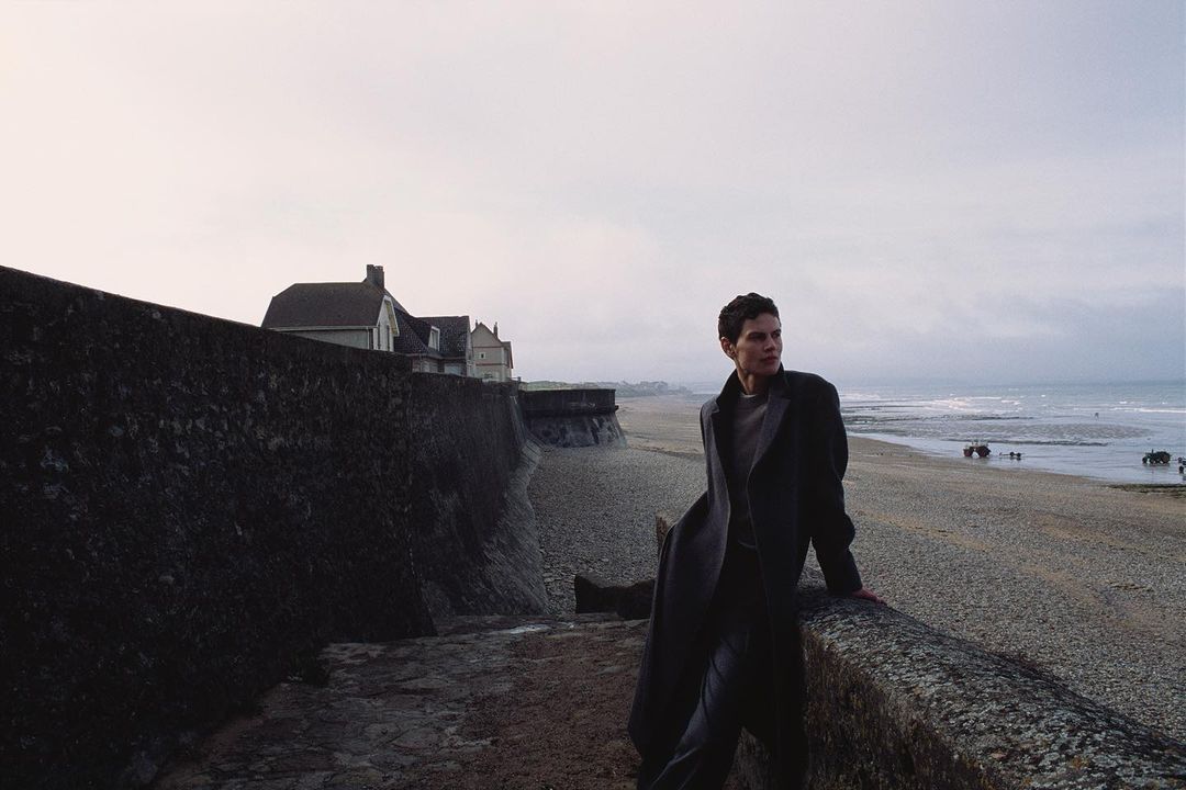 Short-haired woman in a dark coat leaning against a stone wall overlooking a wide sandy beach and sea.