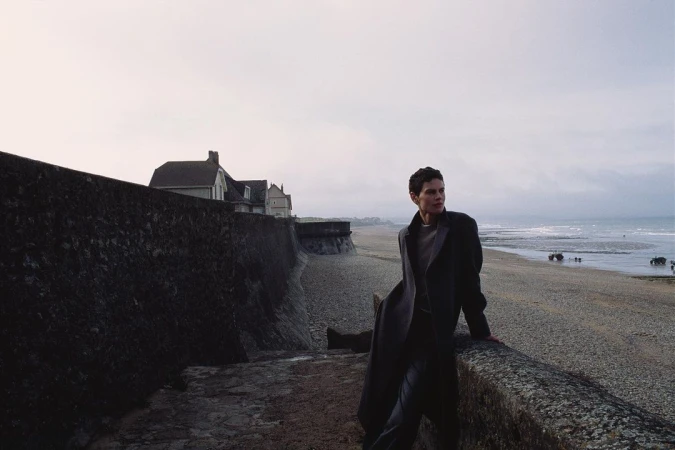 Short-haired woman in a dark coat leaning against a stone wall overlooking a wide sandy beach and sea.