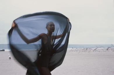 Model with short hair holding a large black sheer fabric on a sandy beach