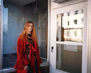 Red-haired female model wearing a red coat standing next to a white door in New York, photographed by Vincent Van De Wijngaard.