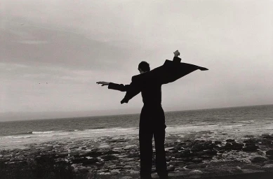 Monochrome rear view of a figure standing on a rocky coast with arms outstretched, coat billowing in the wind against an overcast sky over the ocean.