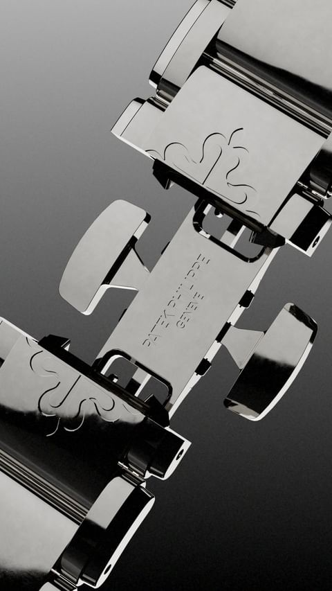 Close-up studio shot of a shiny metal Patek Philippe watch bracelet featuring a deployment clasp with engraved details on a grey gradient background.