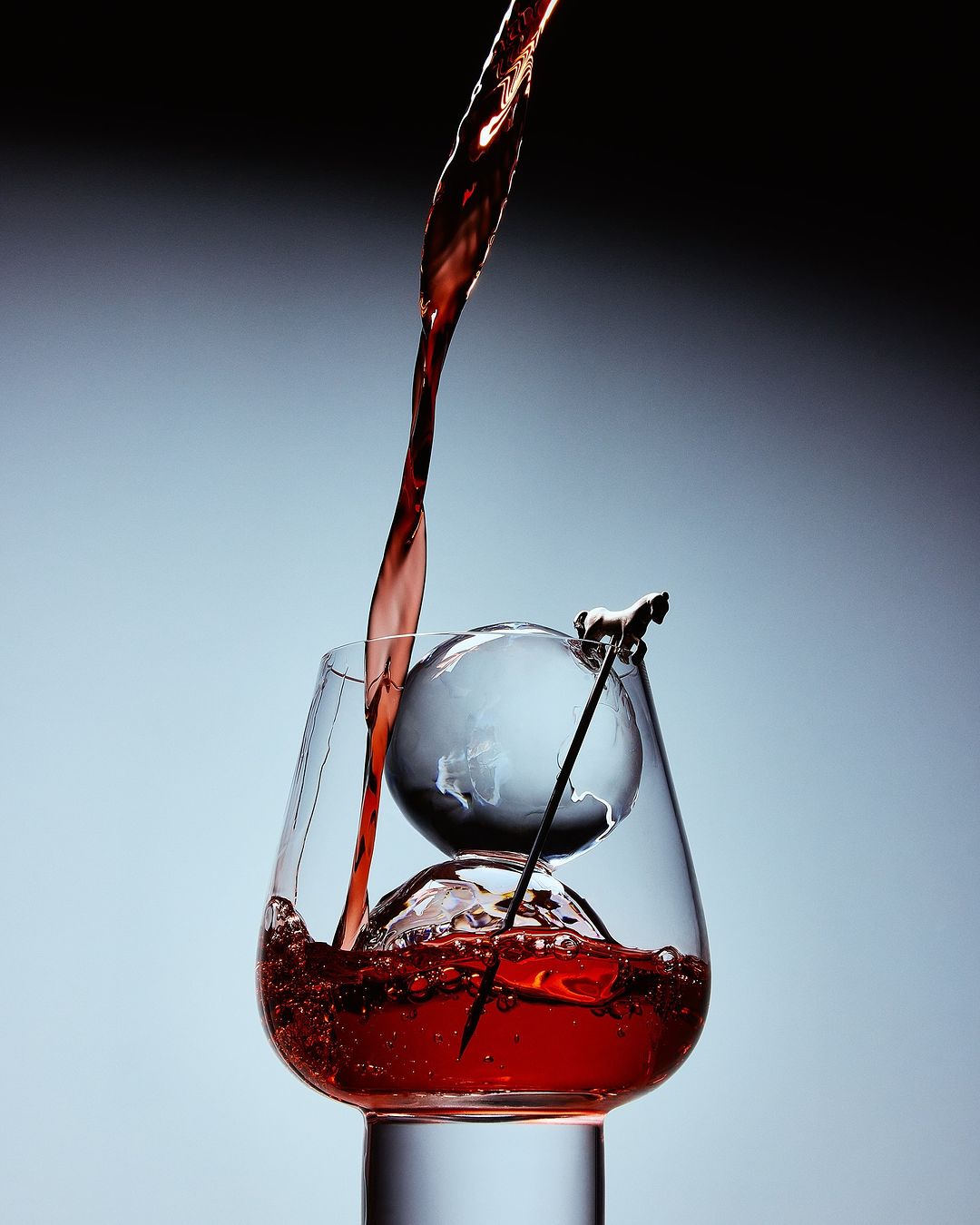 Red liquid pours into a glass containing an ice ball with a globe design, topped with a horse figurine, against a gradient background.