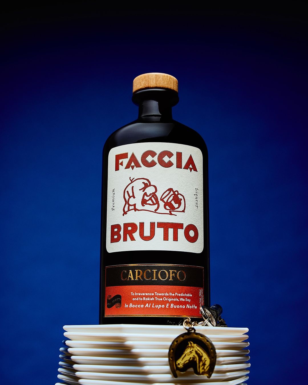 Faccia Brutto artichoke liqueur bottle on a stack of white plates against a graduated blue background. Horseshoe charm hangs from bottle.