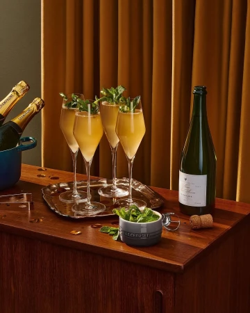 Champagne flutes filled with a golden beverage and garnished with mint sprigs are displayed on a wooden surface, alongside champagne bottles.